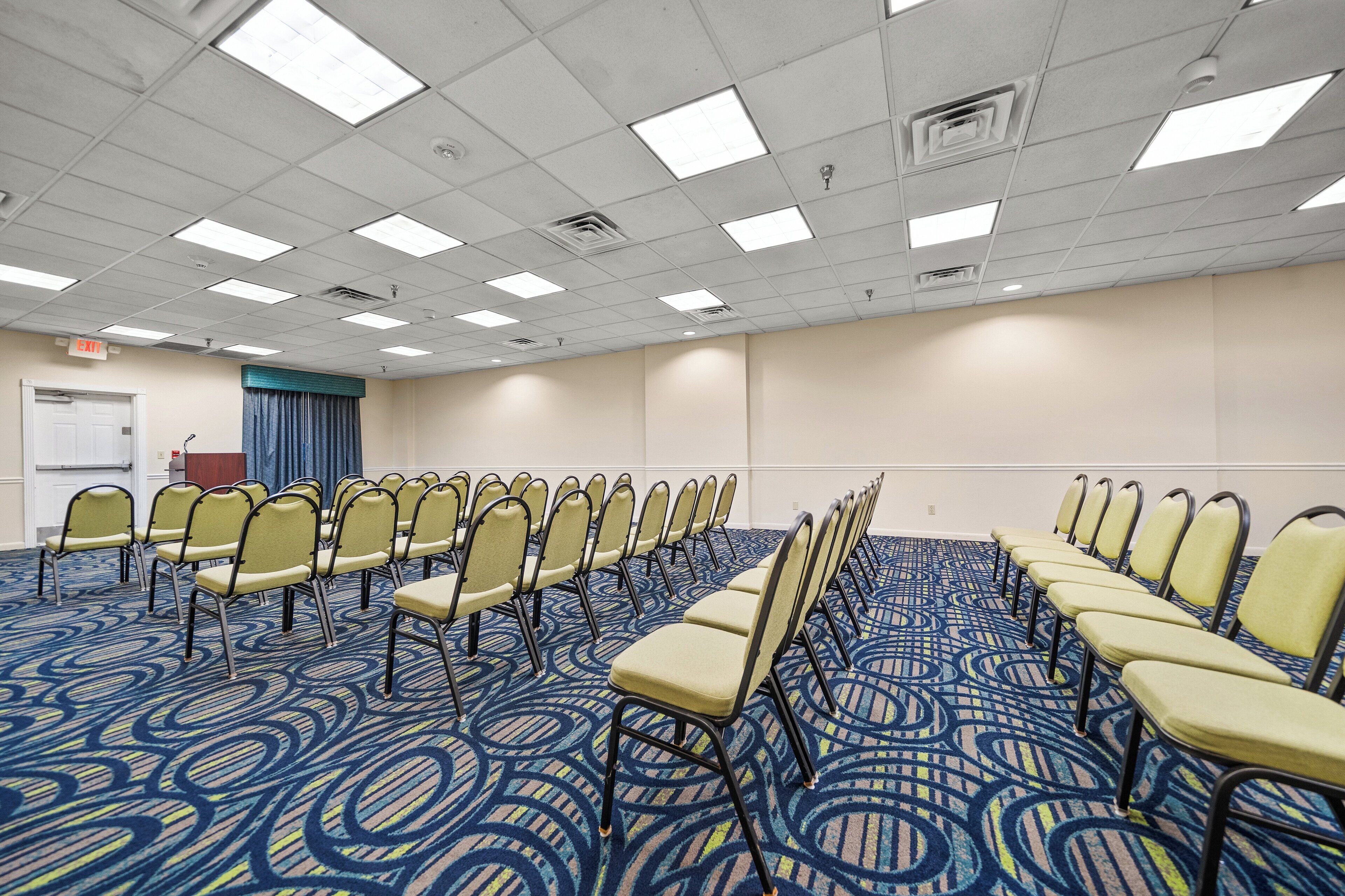 Une salle de conférence avec des rangées de chaises, une moquette à motifs et un plafond blanc équipé de luminaires.