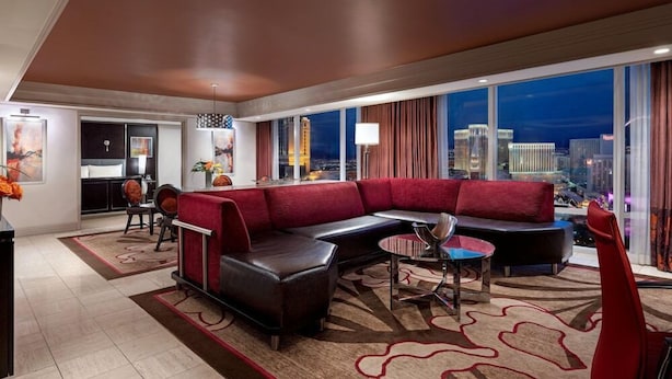 Een moderne hotelkamer met een groot raam met uitzicht op de stad, een rode bank, een eethoek en een aparte zithoek.