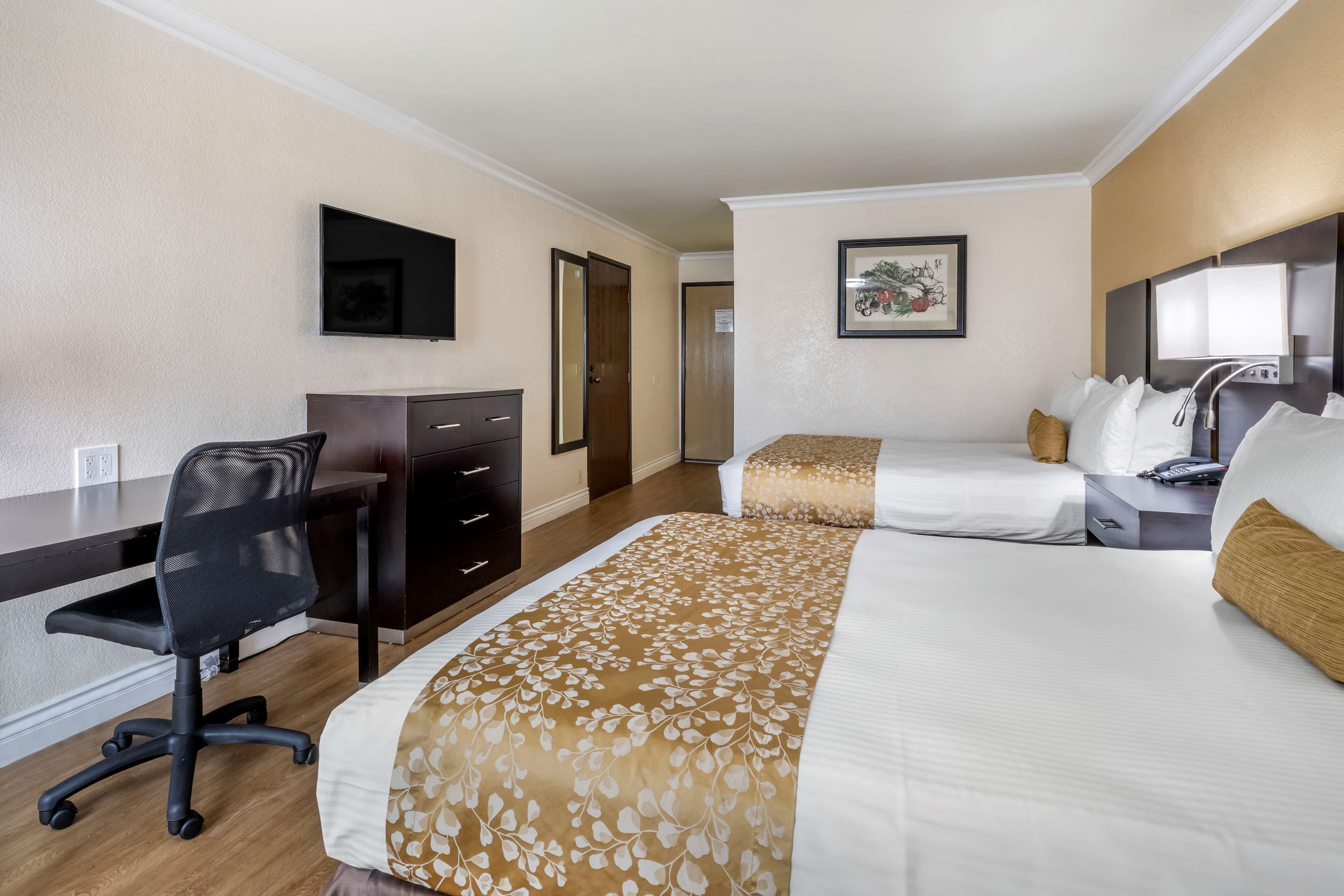 A hotel room with a desk, two beds, a chair, a TV, and a framed picture on the wall.