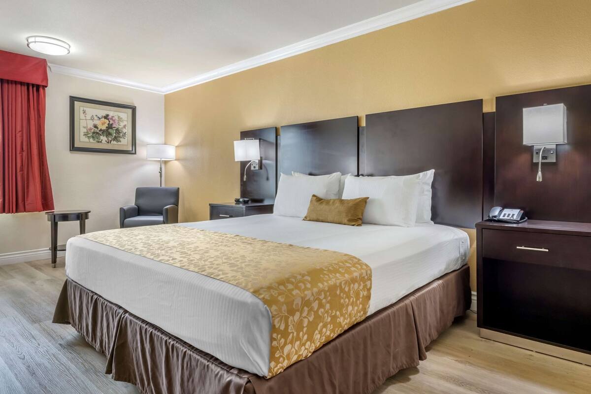 Ett hotellrum med en stor säng, sängbord, ett skrivbord, en stol, en lampa, en telefon och en tavla på väggen.