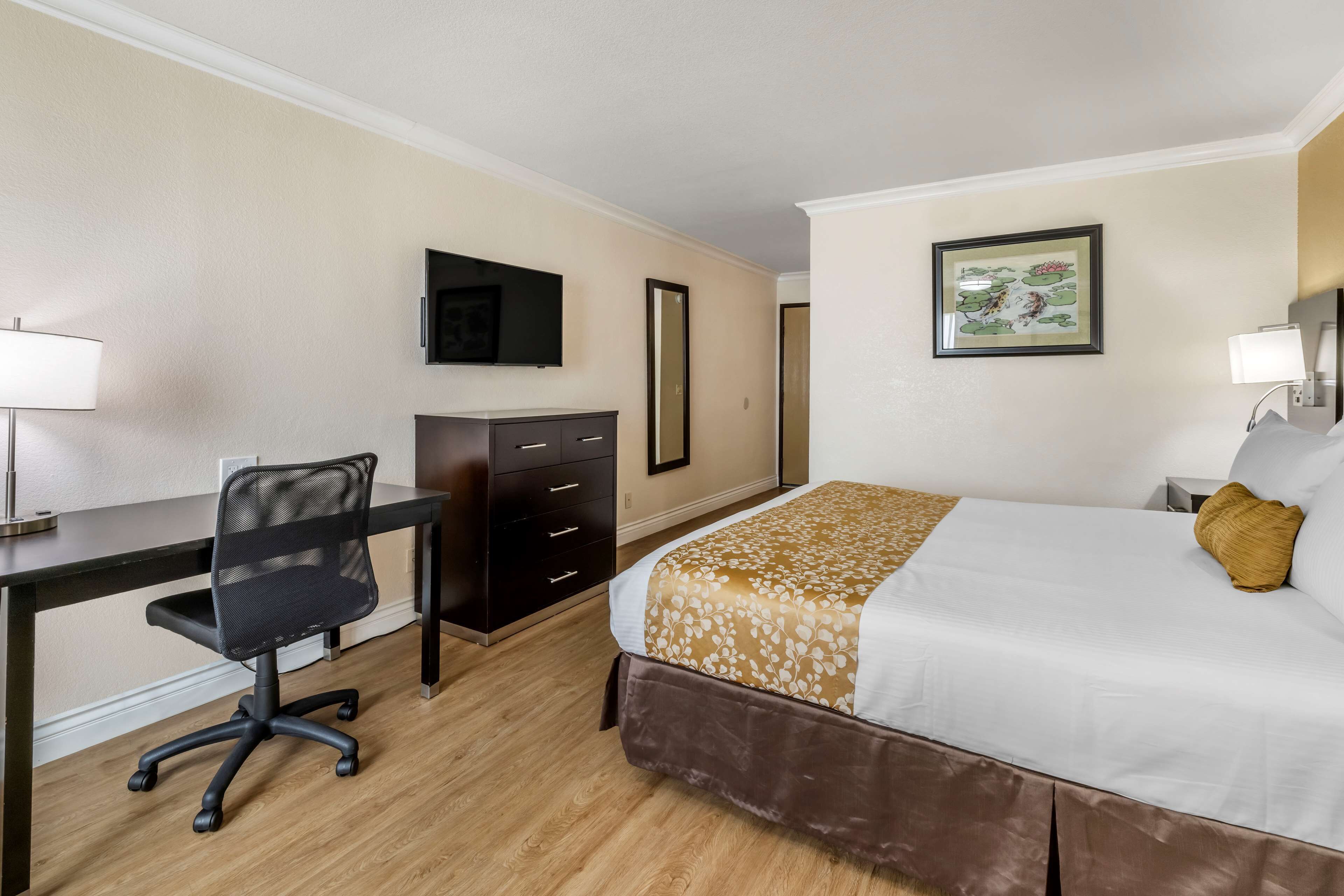 Una camera d'albergo con un letto, una scrivania, una sedia, un comò, una TV e opere d'arte alle pareti.