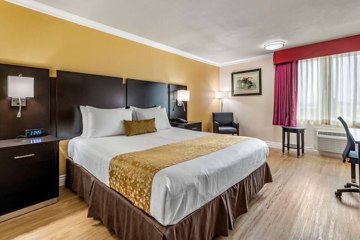 Ett hotellrum med en stor säng, ett skrivbord, en stol, ett nattduksbord, en lampa och ett fönster med gardiner.