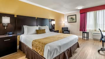 A hotel room with a large bed, a desk, a chair, a nightstand, a lamp, and a window with curtains.