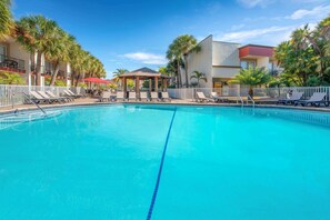 A swimming pool with lounge chairs and a gazebo, surrounded by palm trees and a building with balconies.