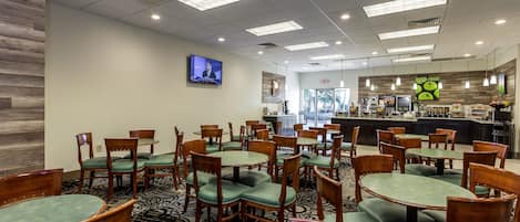 A hotel dining area with multiple round tables and chairs, a television mounted on the wall, and a kitchen area in the background.