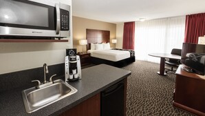 Une chambre d’hôtel équipée d’une kitchenette, comprenant un four à micro-ondes, une machine à café et un évier. La chambre dispose d’un lit avec une tête de lit, d’une télévision et d’un bureau avec une chaise.