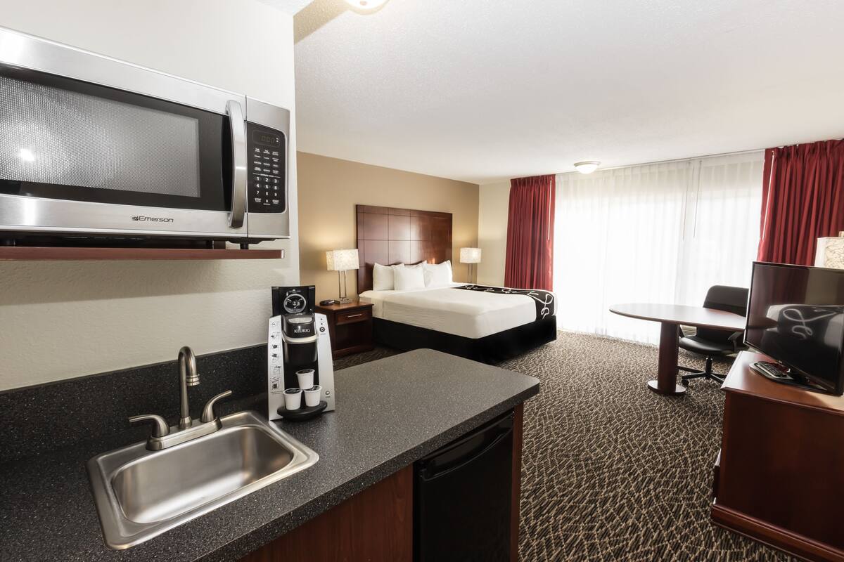 Habitación de hotel con kitchenette, que incluye microondas, cafetera y lavabo. Cuenta con una cama con cabecera, un televisor y un escritorio con silla.
