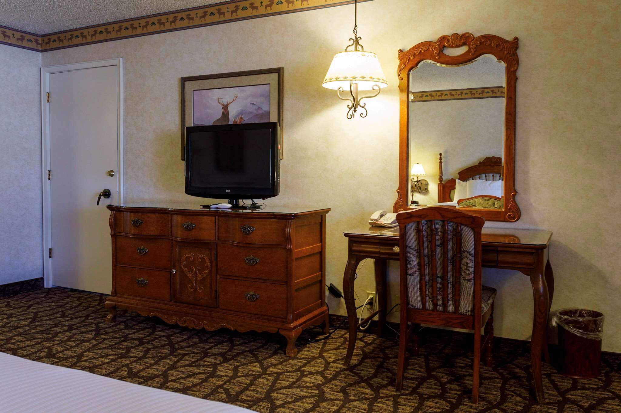 A hotel room with a wooden dresser, a television, a desk with a chair, a mirror, and a patterned carpet.