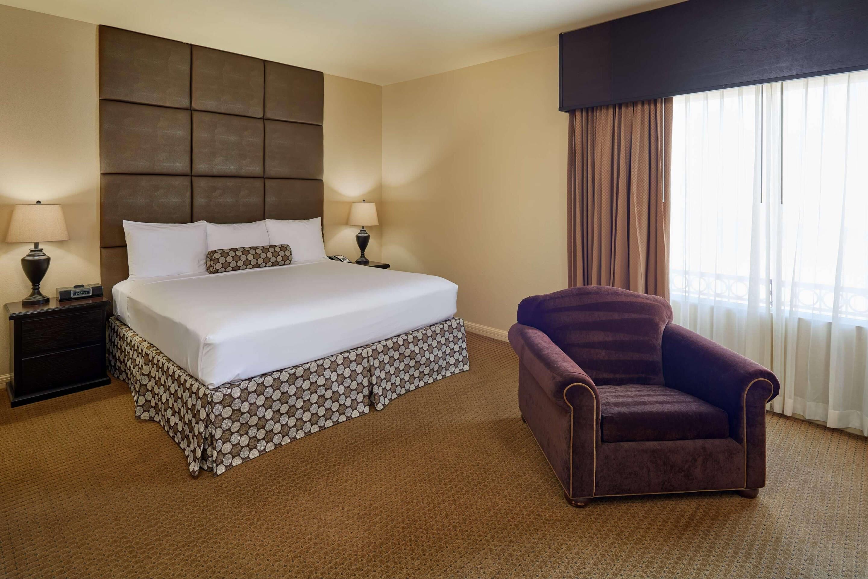 A hotel room with a large bed, two bedside tables with lamps, a purple armchair, and a window with curtains.