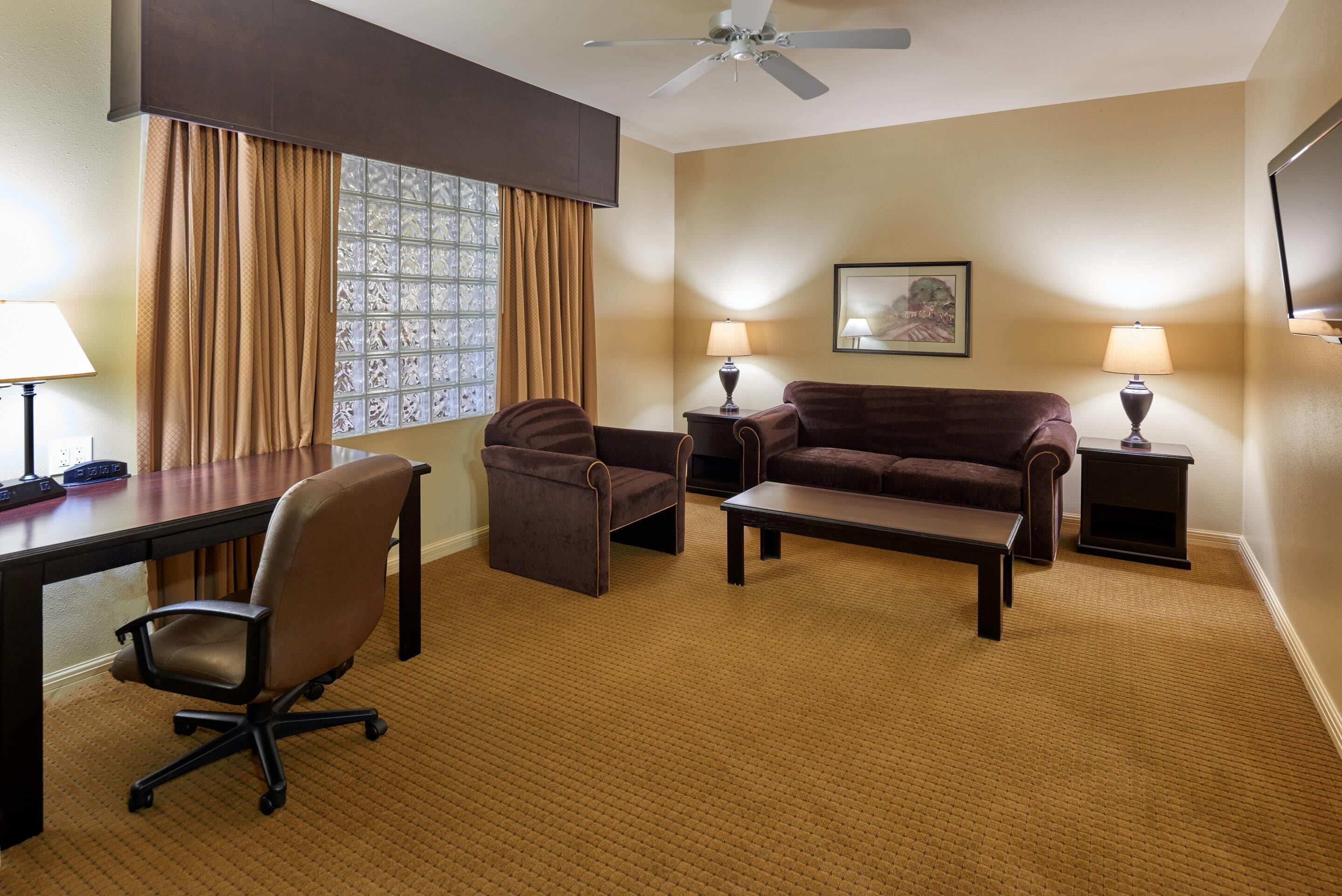 Habitación de hotel con escritorio, silla de oficina, dos sofás, una mesa de centro, lámparas, ventilador de techo y una ventana con cortinas.