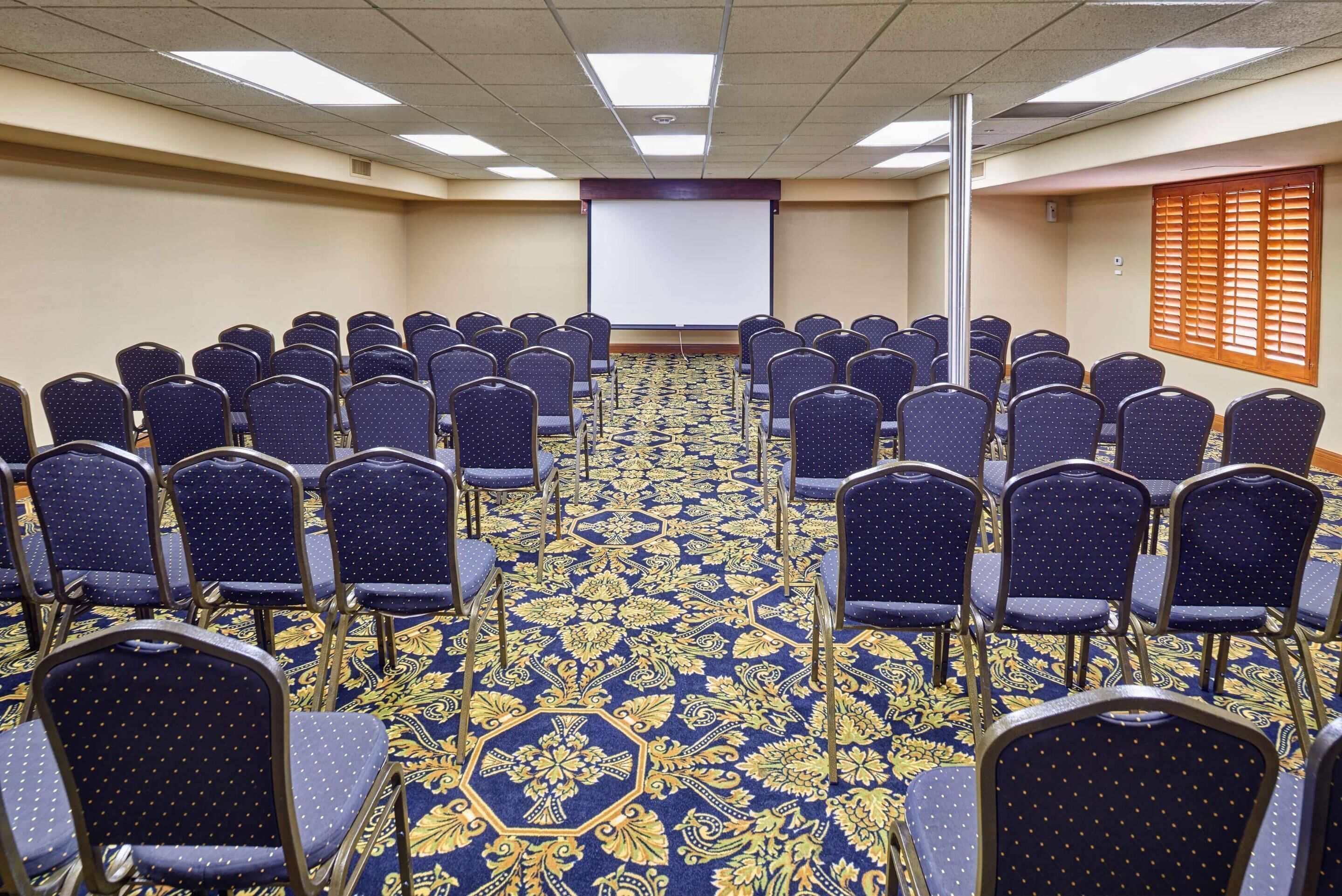 A conference room with rows of chairs facing a projector screen, carpeted floor, and a window with blinds.