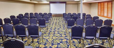 A conference room with rows of chairs facing a projector screen, carpeted floor, and a window with blinds.
