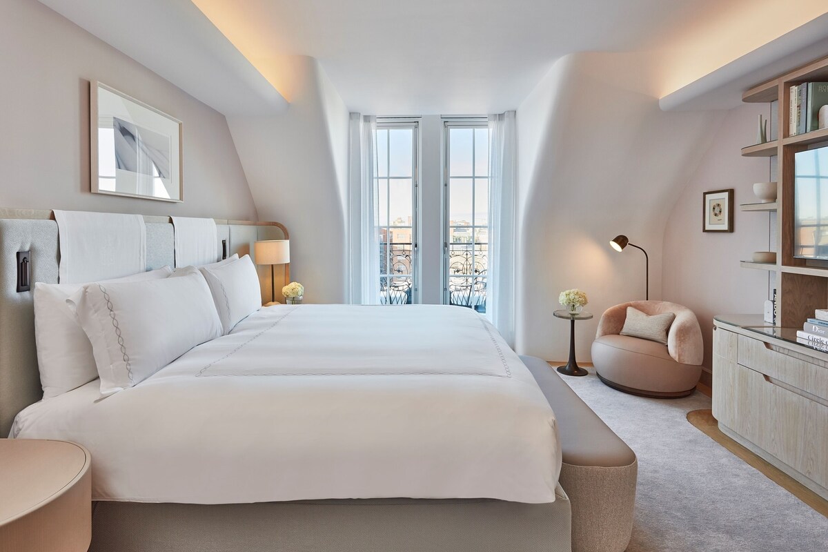 A modern hotel room with a large bed, a round chair, a small table, and a bookshelf.