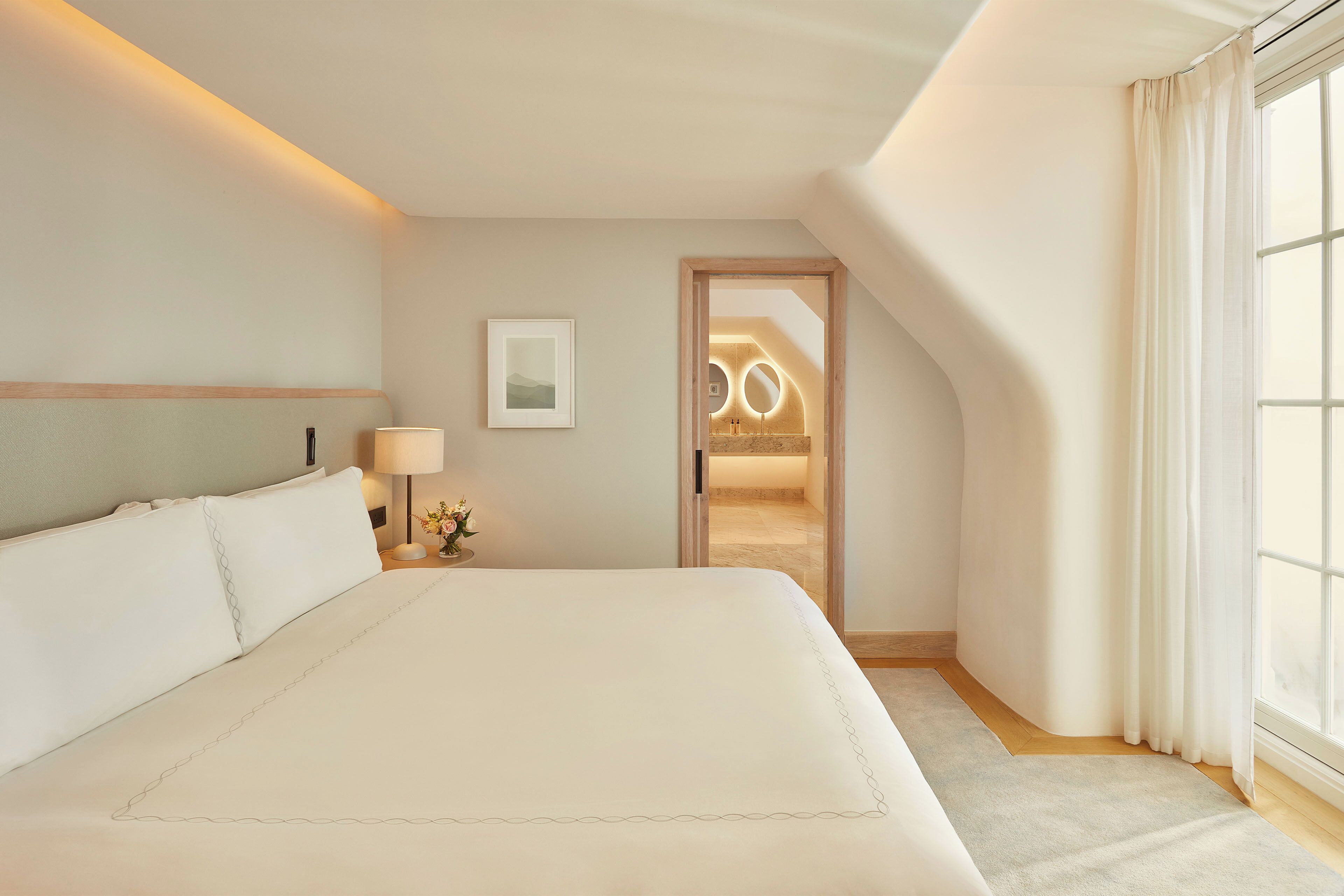 Ein großes Bett mit Kopfteil, ein Nachttisch mit Lampe, ein gerahmtes Bild an der Wand und Blick auf ein Badezimmer mit Spiegel und runder Leuchte.