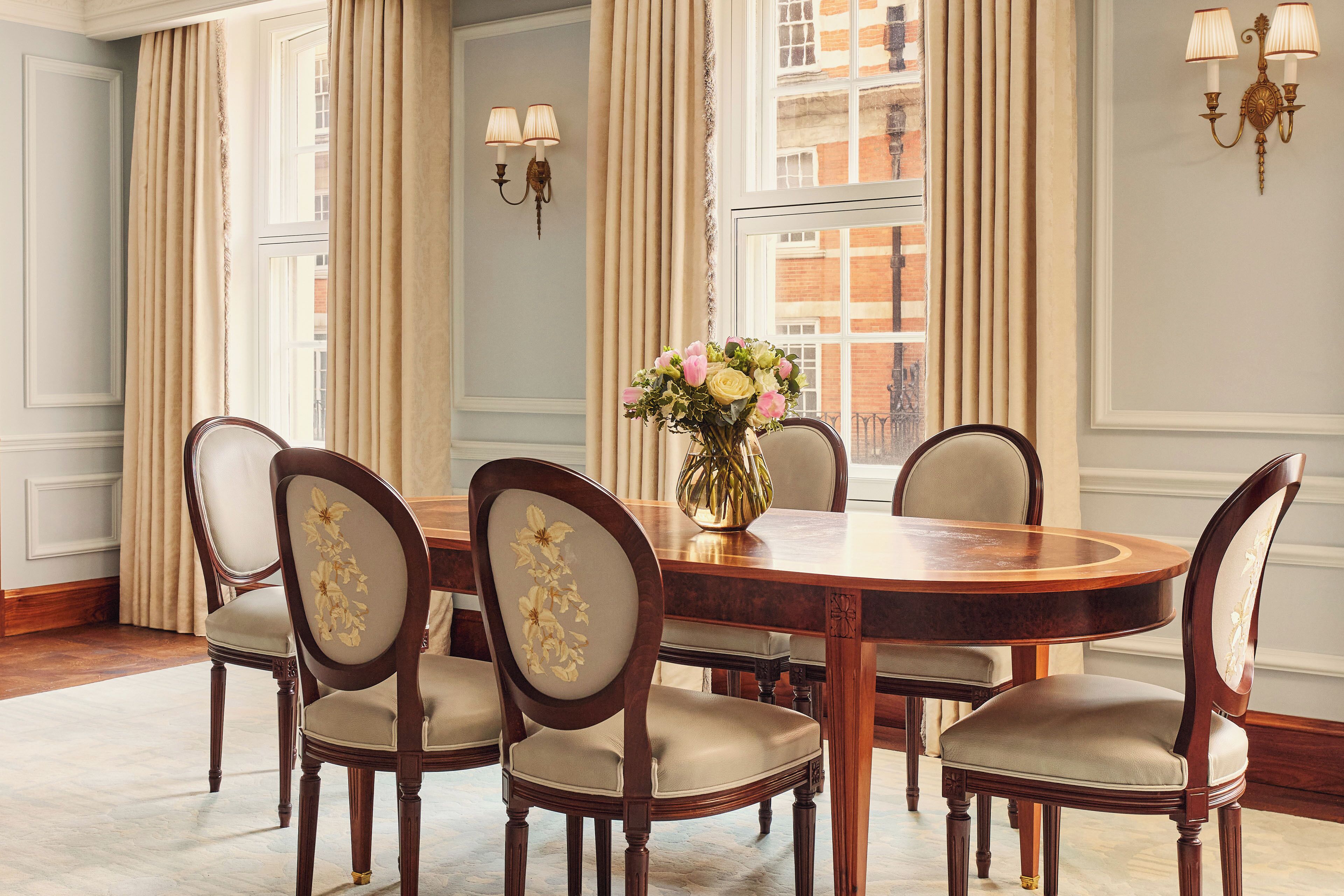 A dining room with a round wooden table, six upholstered chairs with floral patterns, a vase of flowers, and a view of a building through large windows.