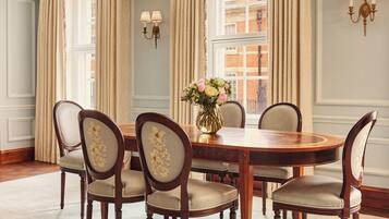 Comedor con una mesa de madera redonda, seis sillas tapizadas con estampados florales, un jarrón de flores y vistas a un edificio a través de amplios ventanales.