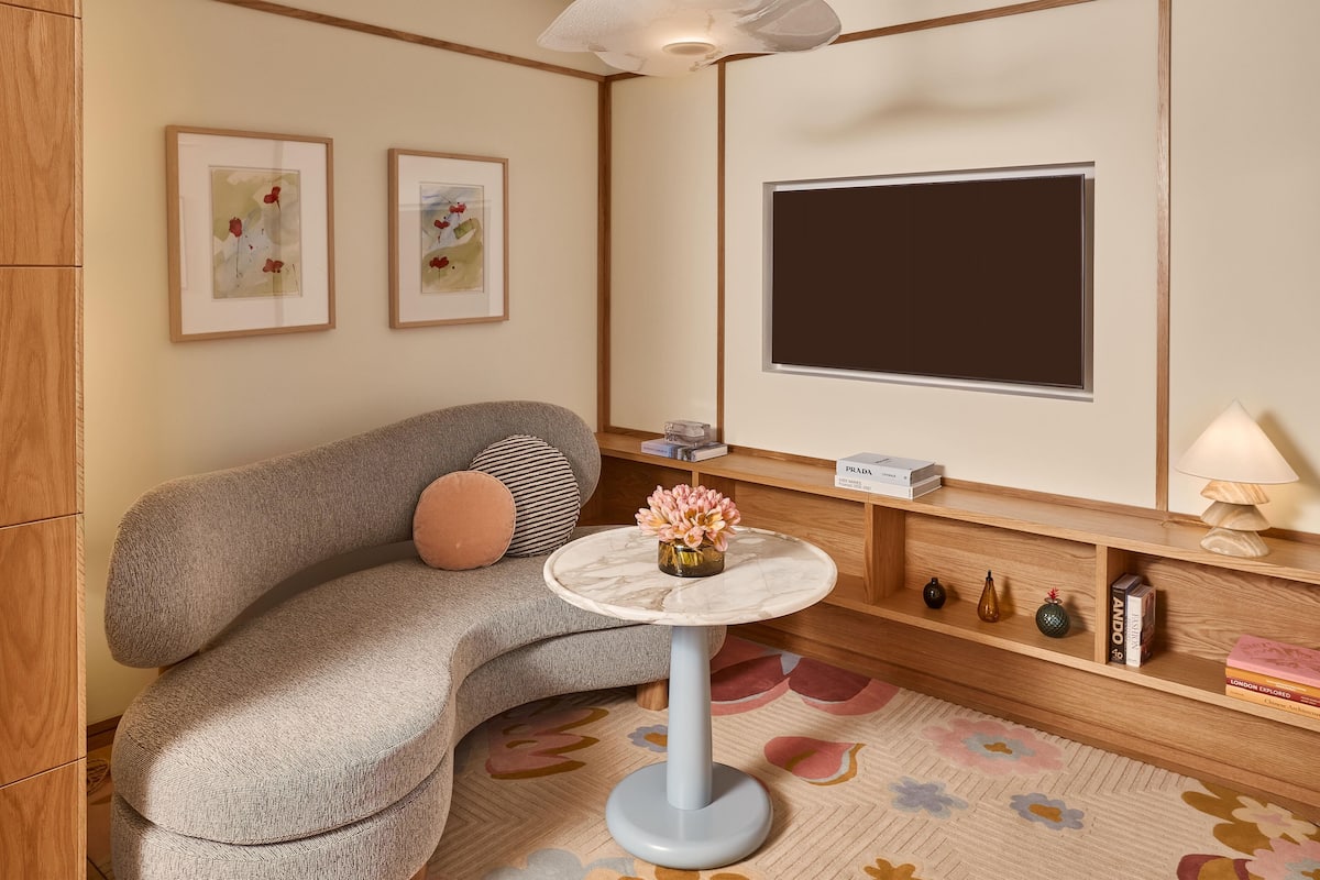 A modern living room with a grey sofa, a round table with a vase of flowers, a flat-screen TV, and a wooden bookshelf.