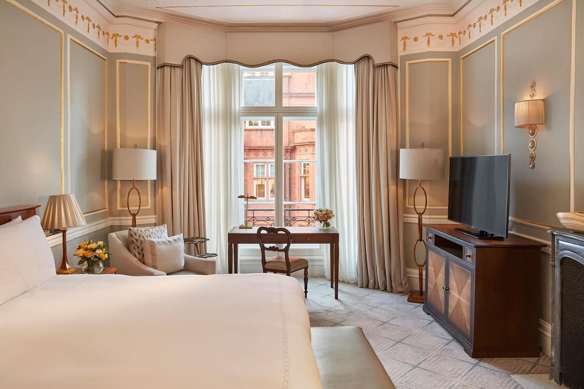 A hotel room with a large bed, a desk with a chair, a television, and a fireplace.