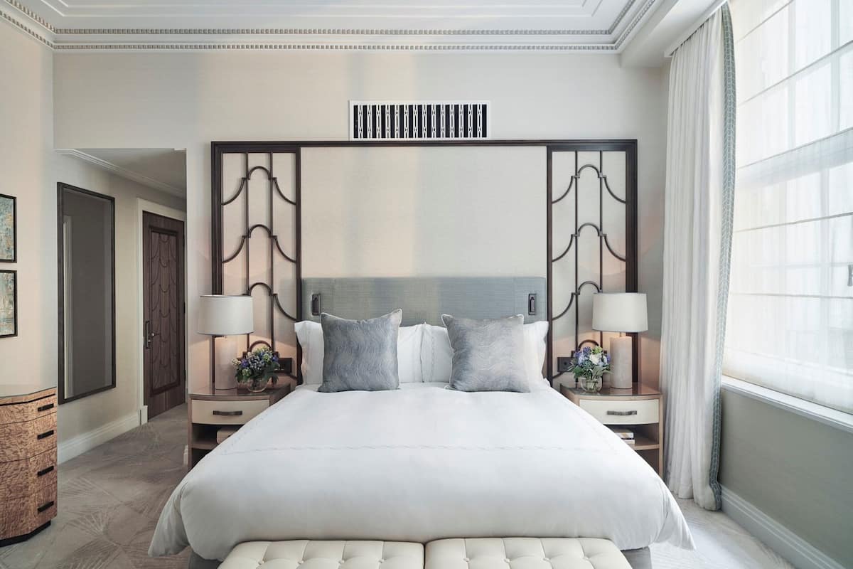 A spacious hotel room with a large bed, two bedside tables, a headboard with decorative panels, a bench at the foot of the bed, and a large window with curtains.