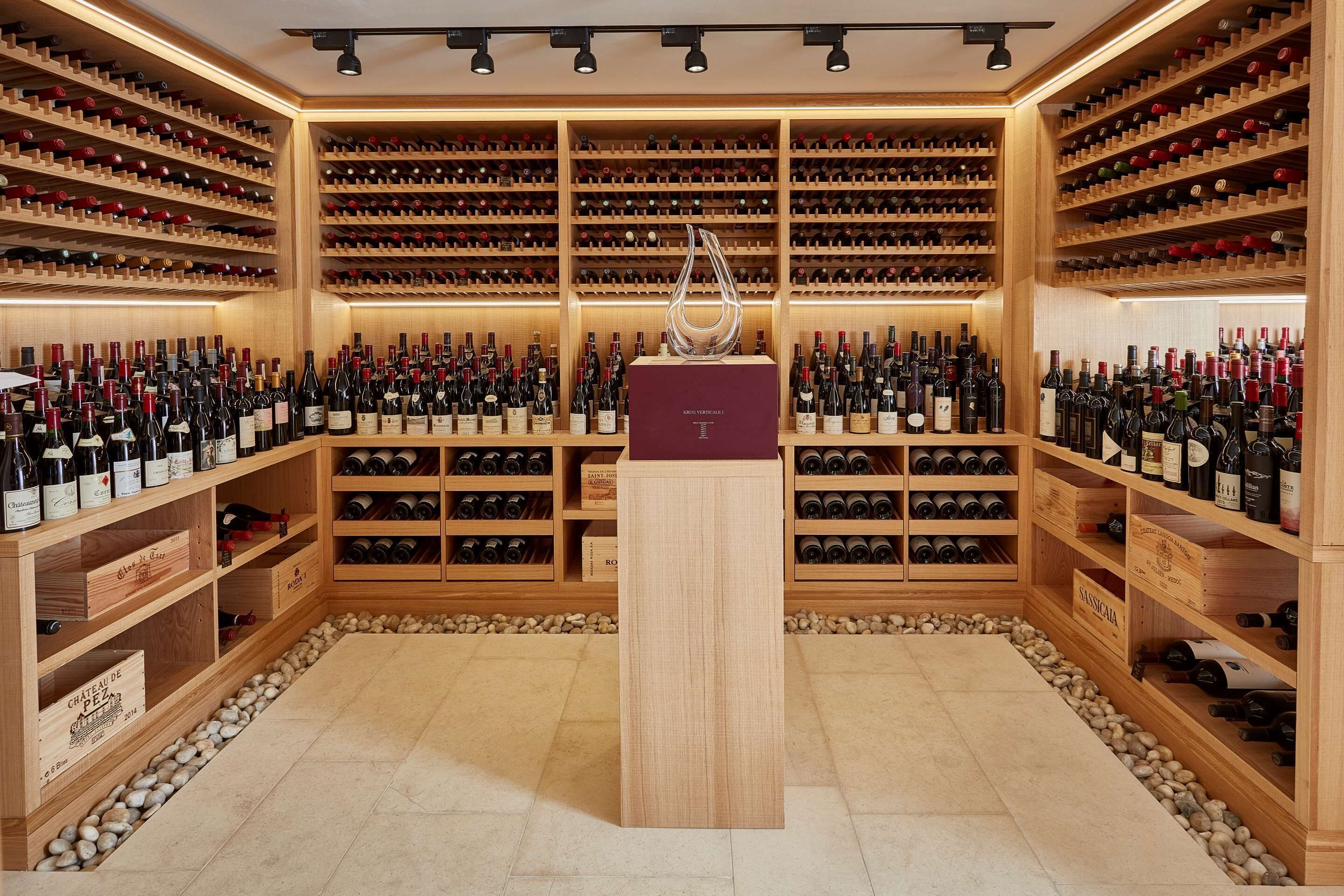 A wine cellar with wooden shelves filled with wine bottles, a central display case, and a stone-tiled floor.