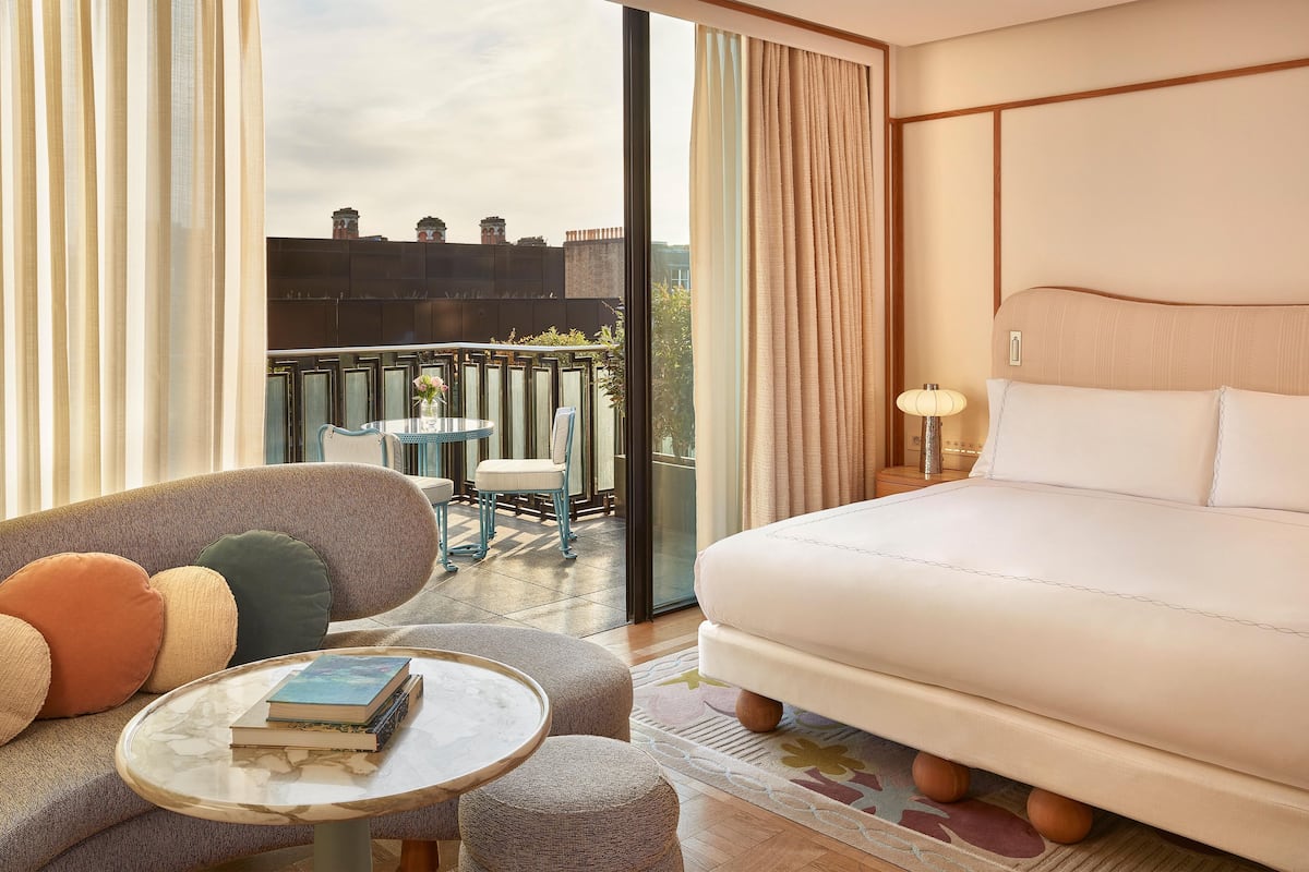 A hotel room with a large bed, a sofa, a small table, and a balcony with a view of buildings.