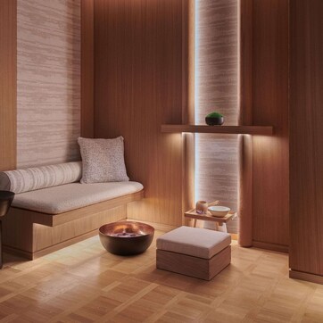 A modern, minimalist room with a wooden floor, a cushioned bench, a small table with a bowl, and a shelf with a plant.