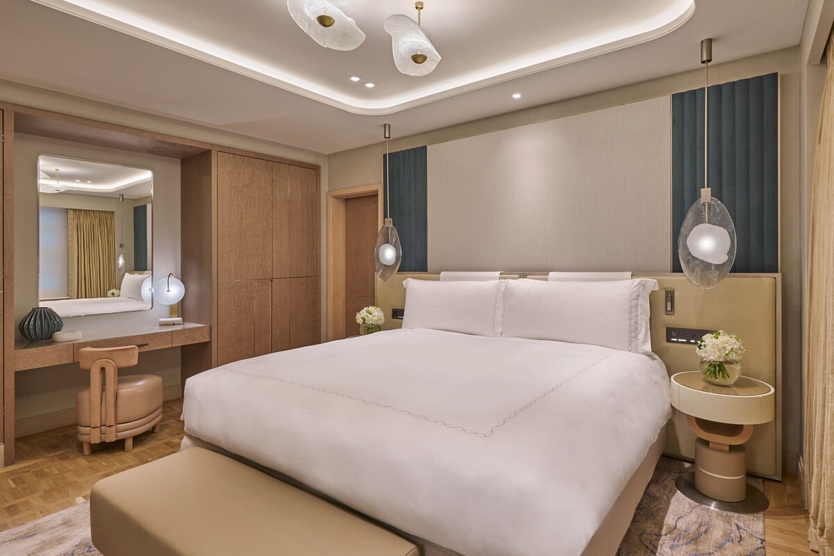 A modern hotel room with a large bed, a vanity with a mirror, a desk, and a large window with curtains.