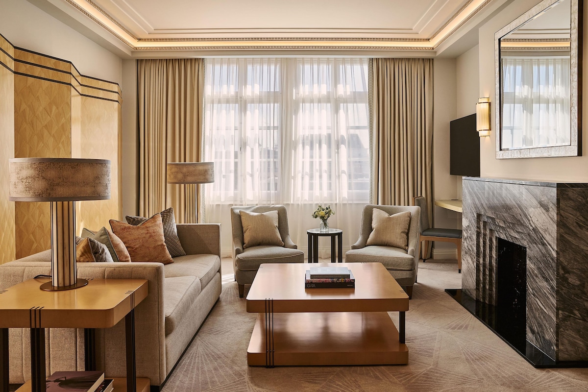 A modern living room with a sofa, two armchairs, a coffee table, a side table, a lamp, a fireplace, and a large window with curtains.