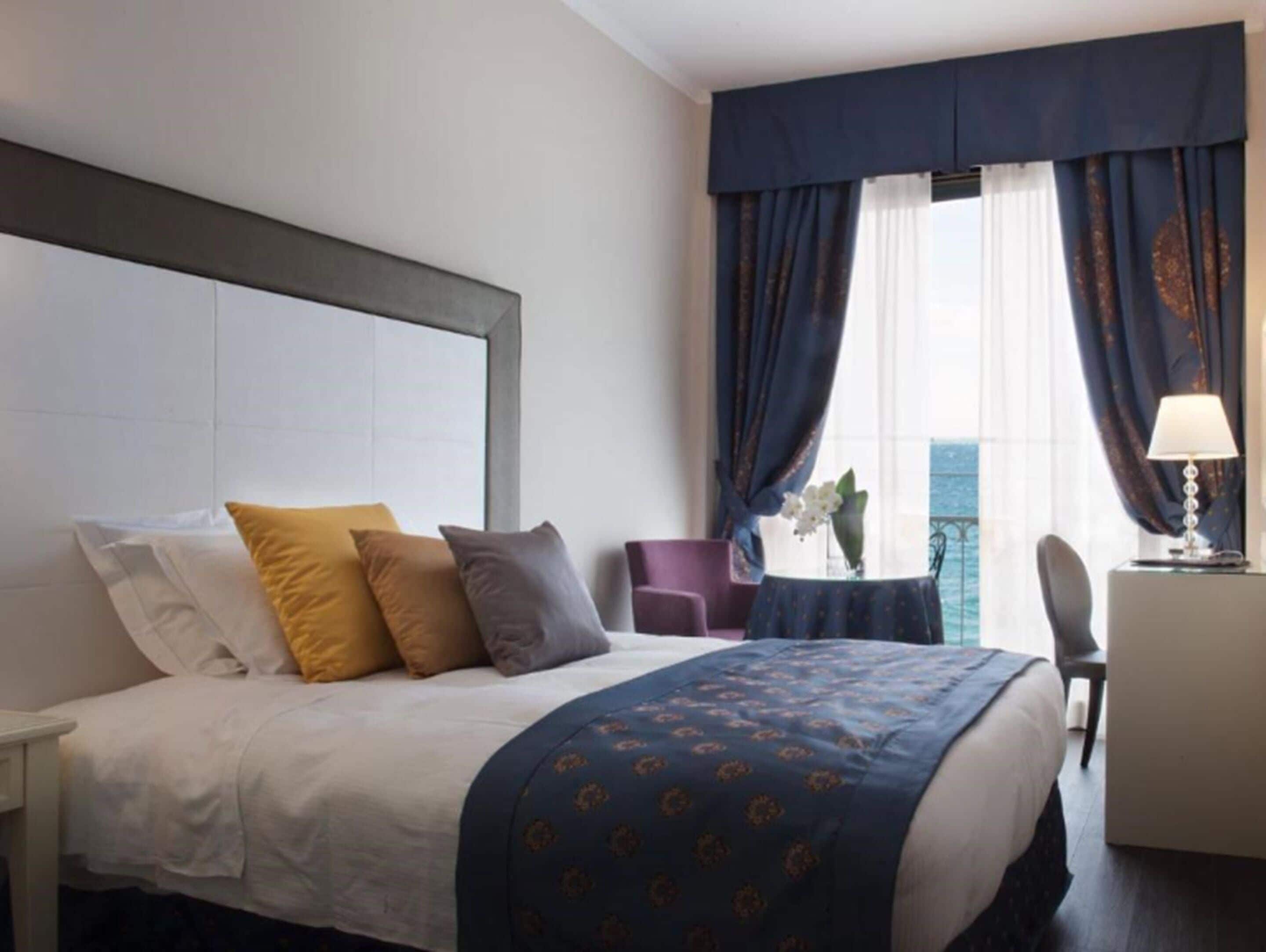 A hotel room with a bed, a desk, a chair, and a view of the sea through large windows with curtains.