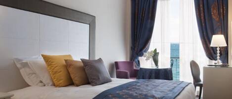 A hotel room with a bed, a desk, a chair, and a view of the sea through large windows with curtains.