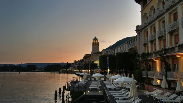 A waterfront hotel with outdoor seating and a view of a tower in the background. - Grand Hotel Gardone Riviera (Gardone Riviera)