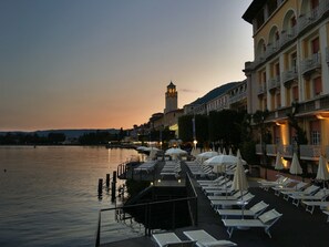 A waterfront hotel with outdoor seating and a view of a tower in the background. - Grand Hotel Gardone Riviera (Gardone Riviera)