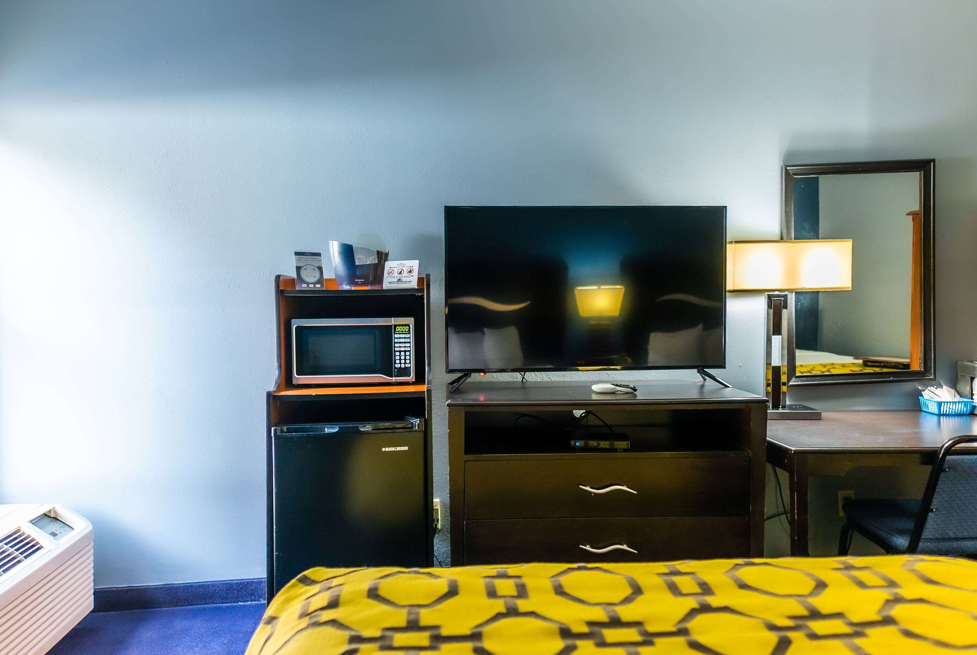 A hotel room with a bed, a desk with a computer, a microwave, a small refrigerator, a TV, and a lamp.