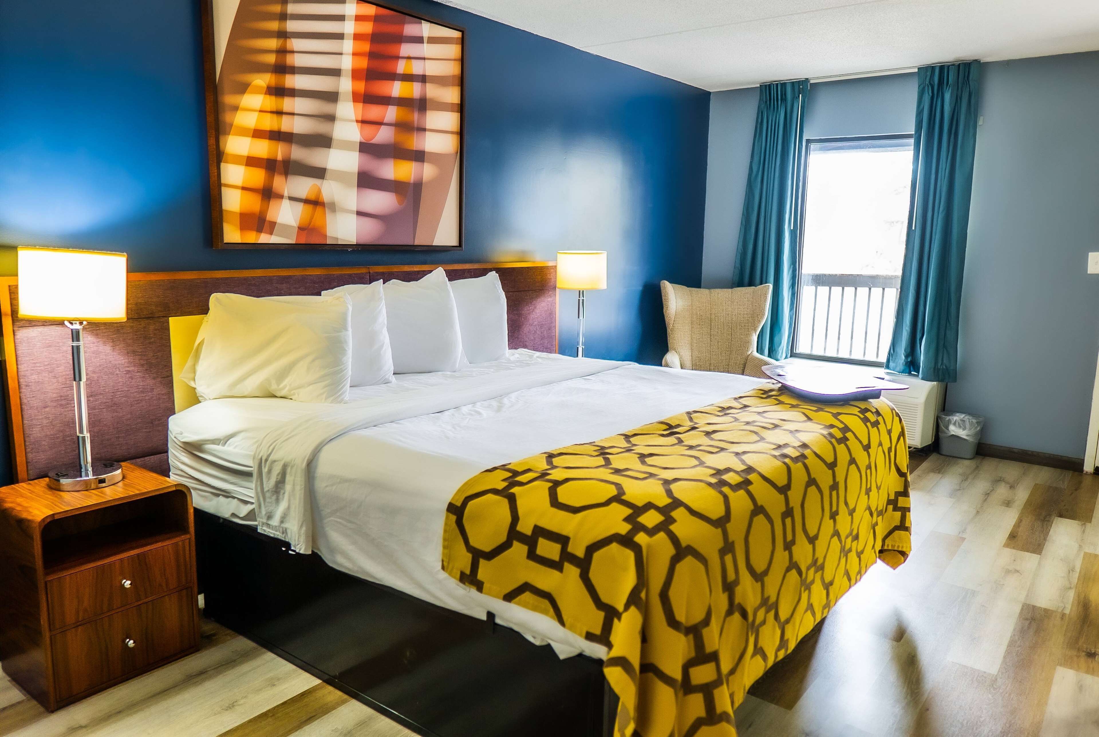 A hotel room with a bed, two bedside lamps, a nightstand, a chair, a window with curtains, and a large abstract painting on the wall.