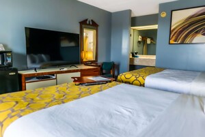 Habitación de hotel con dos camas, un escritorio, televisor, espejo y baño visible a través de una puerta abierta.