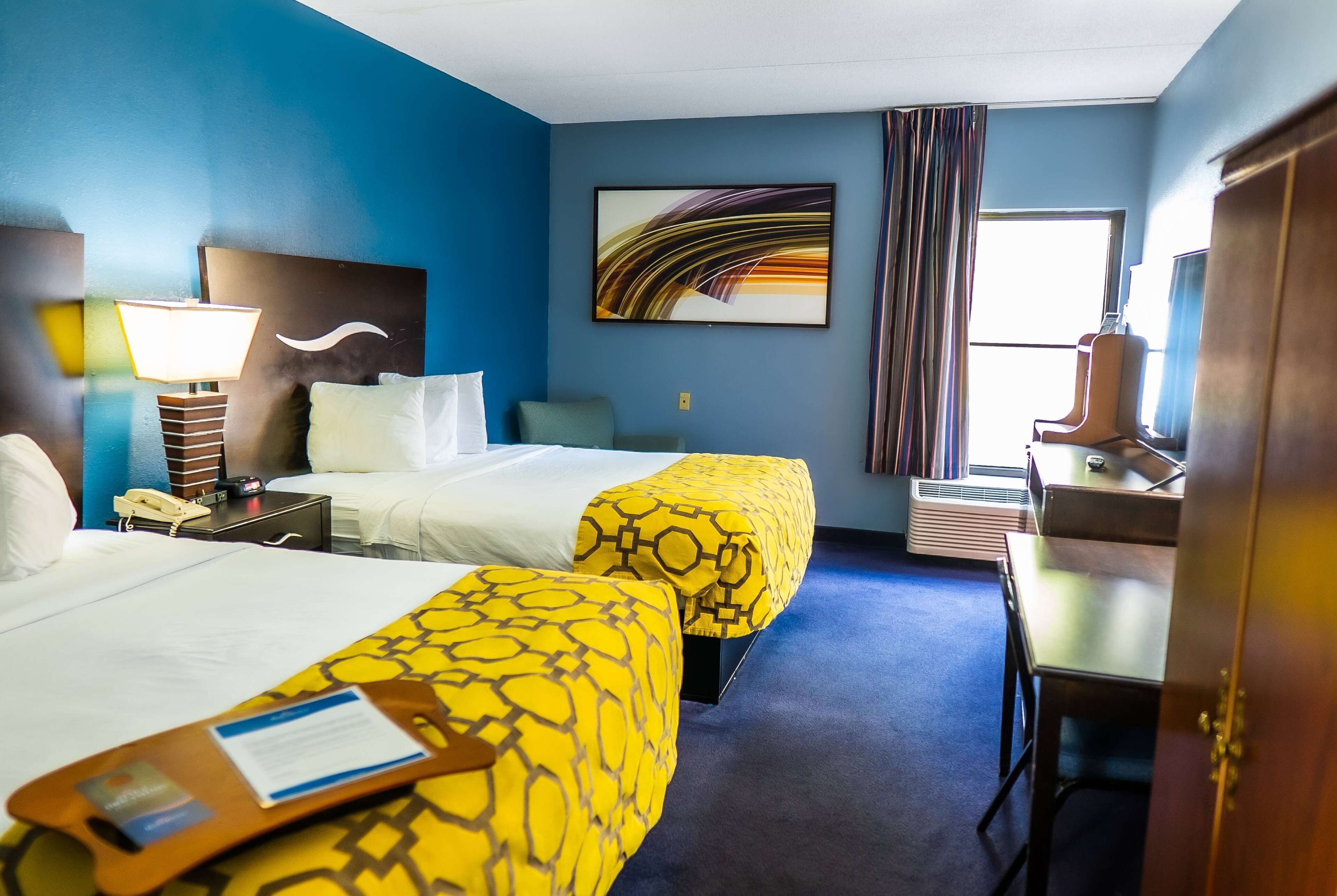 A hotel room with two beds, a desk, a lamp, a painting, and a window with curtains.