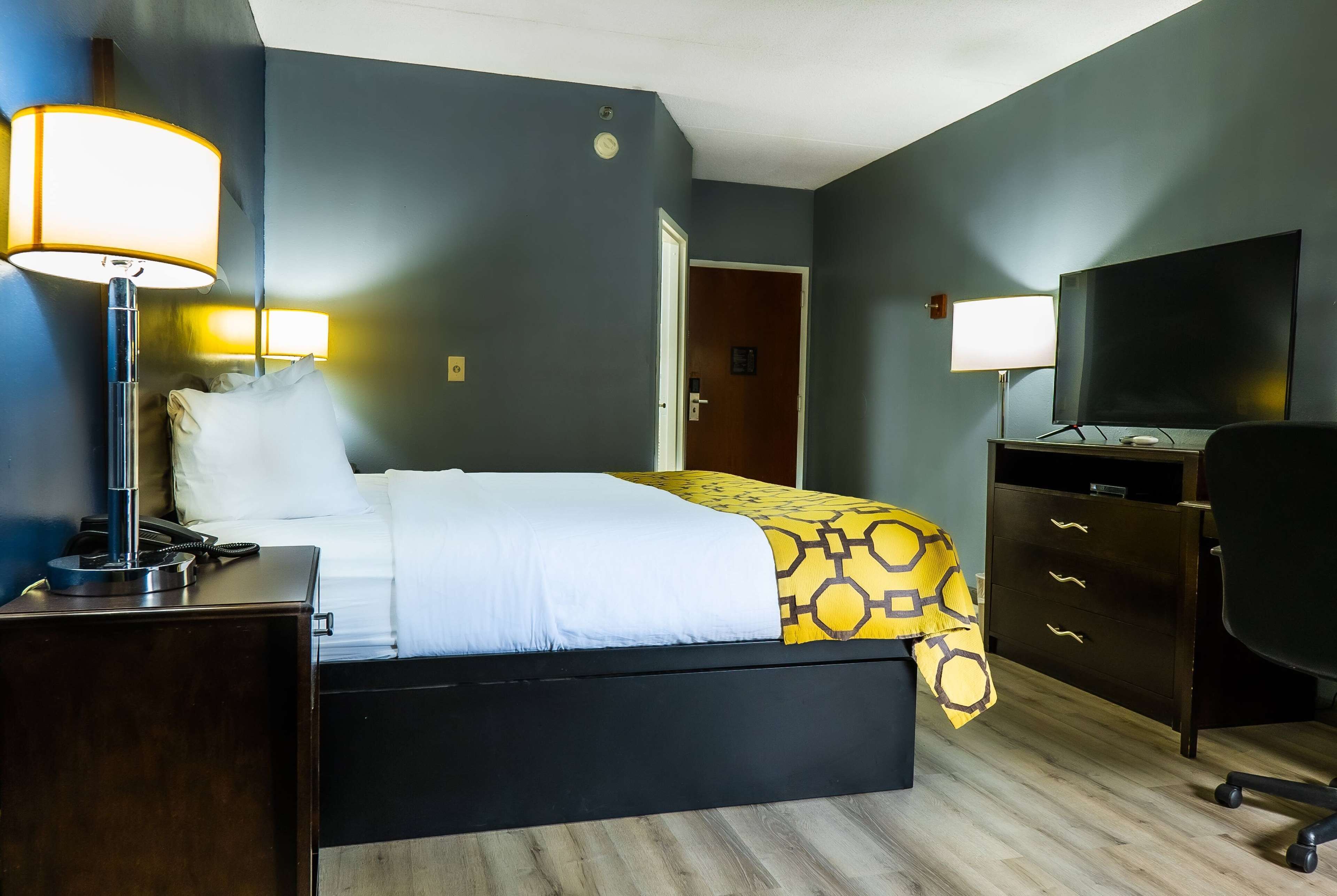 Habitación de hotel con una cama, lámparas de noche, un escritorio con teléfono, cómoda, televisor y una puerta que da a otra habitación.