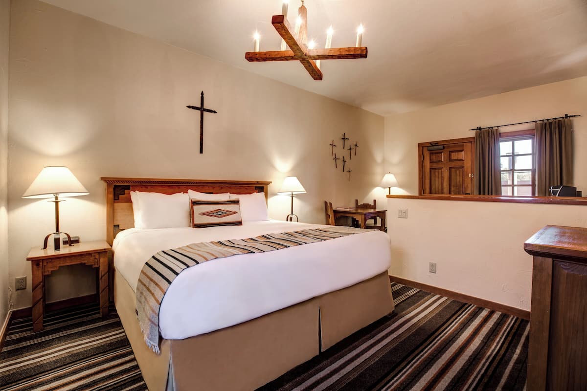 A hotel room with a large bed, two bedside tables with lamps, a wooden headboard, a cross on the wall, and a window with curtains.