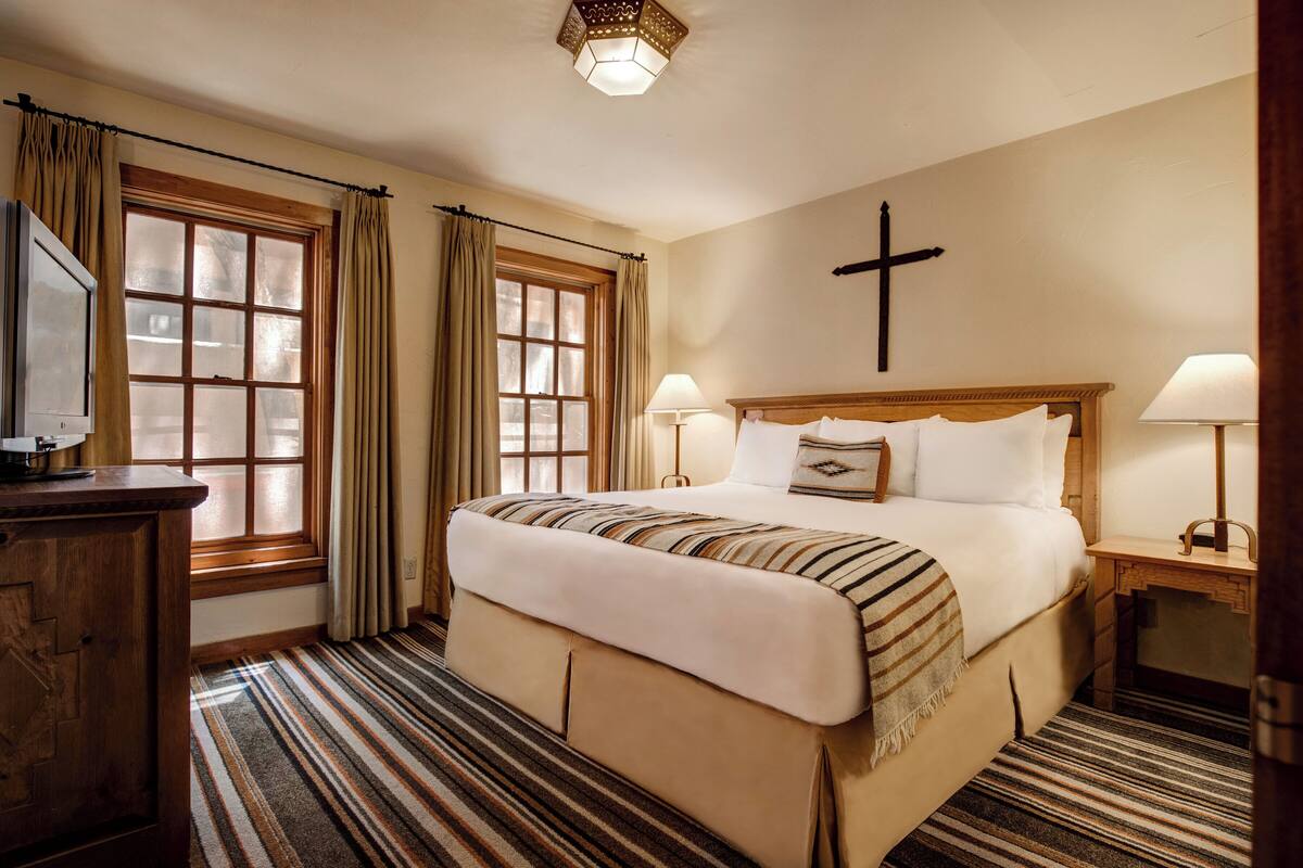 A hotel room with a large bed, two bedside lamps, a wooden headboard, a cross on the wall, and a window with curtains.