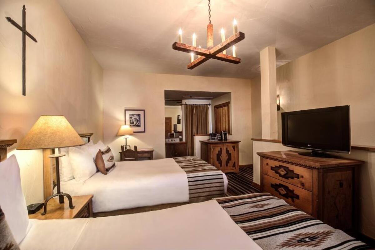 A hotel room with two beds, a television, a wooden cabinet, a cross on the wall, and a chandelier.
