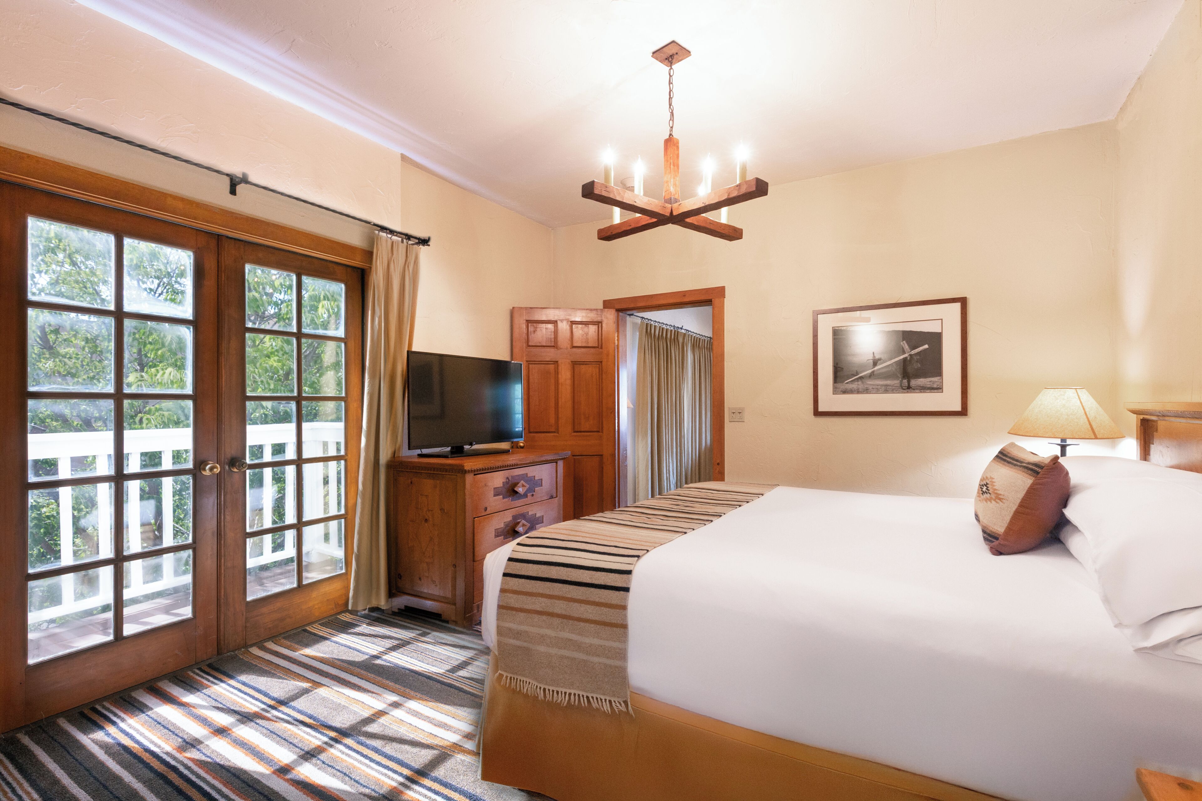 A hotel room with a bed, a television, a wooden dresser, a chandelier, and a large window with curtains.