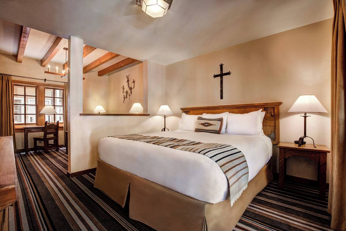 A neatly arranged hotel room with a large bed, bedside lamps, a wooden headboard, a cross on the wall, and a window with curtains.