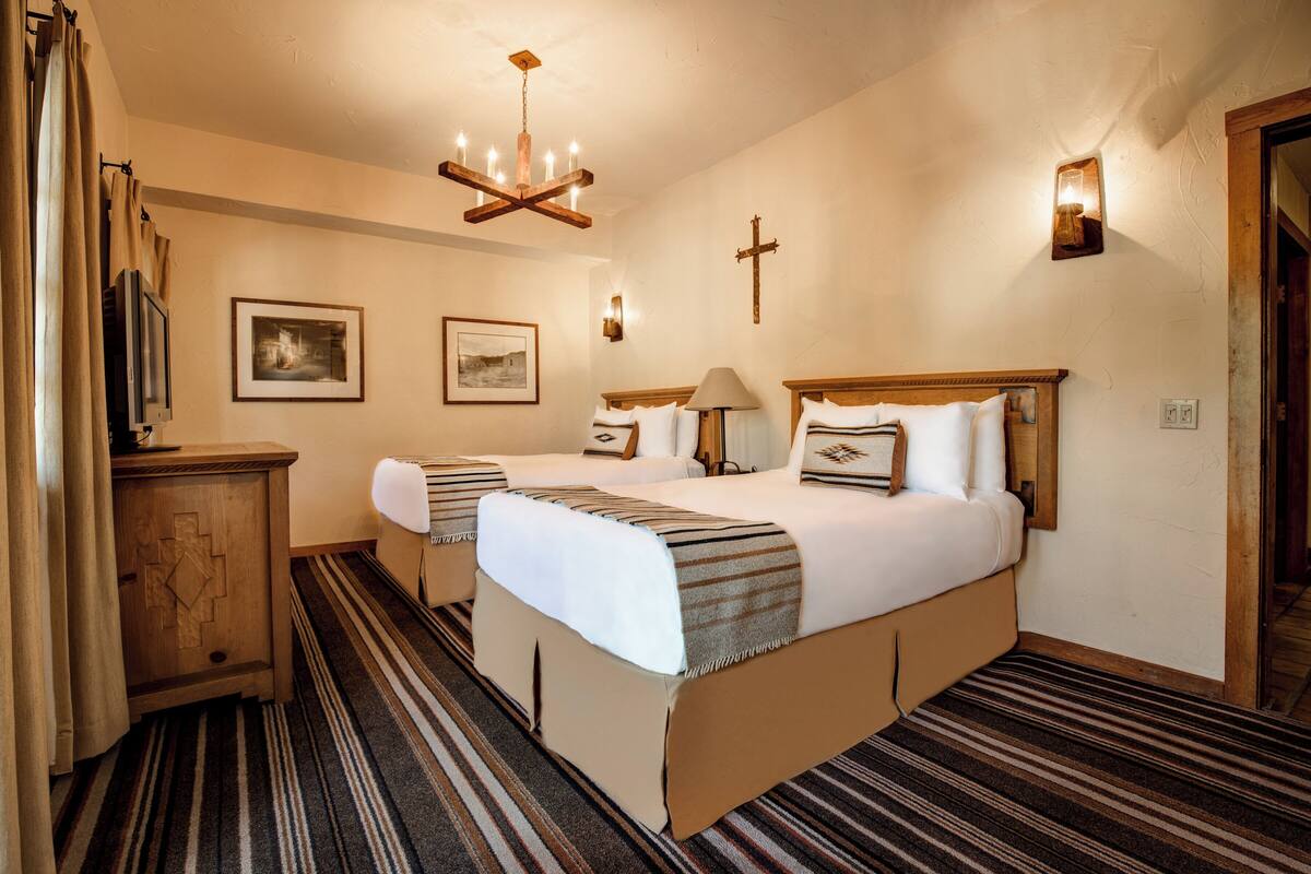 A hotel room with two beds, a wooden headboard, a chandelier, a television, and a framed picture on the wall.