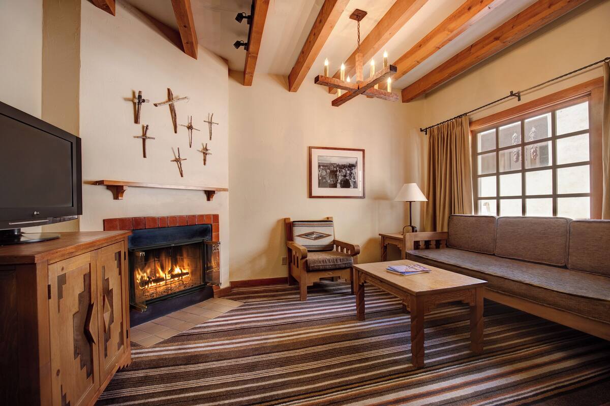 A cozy living room with a fireplace, wooden beams, a sofa, a TV, and a patterned rug.