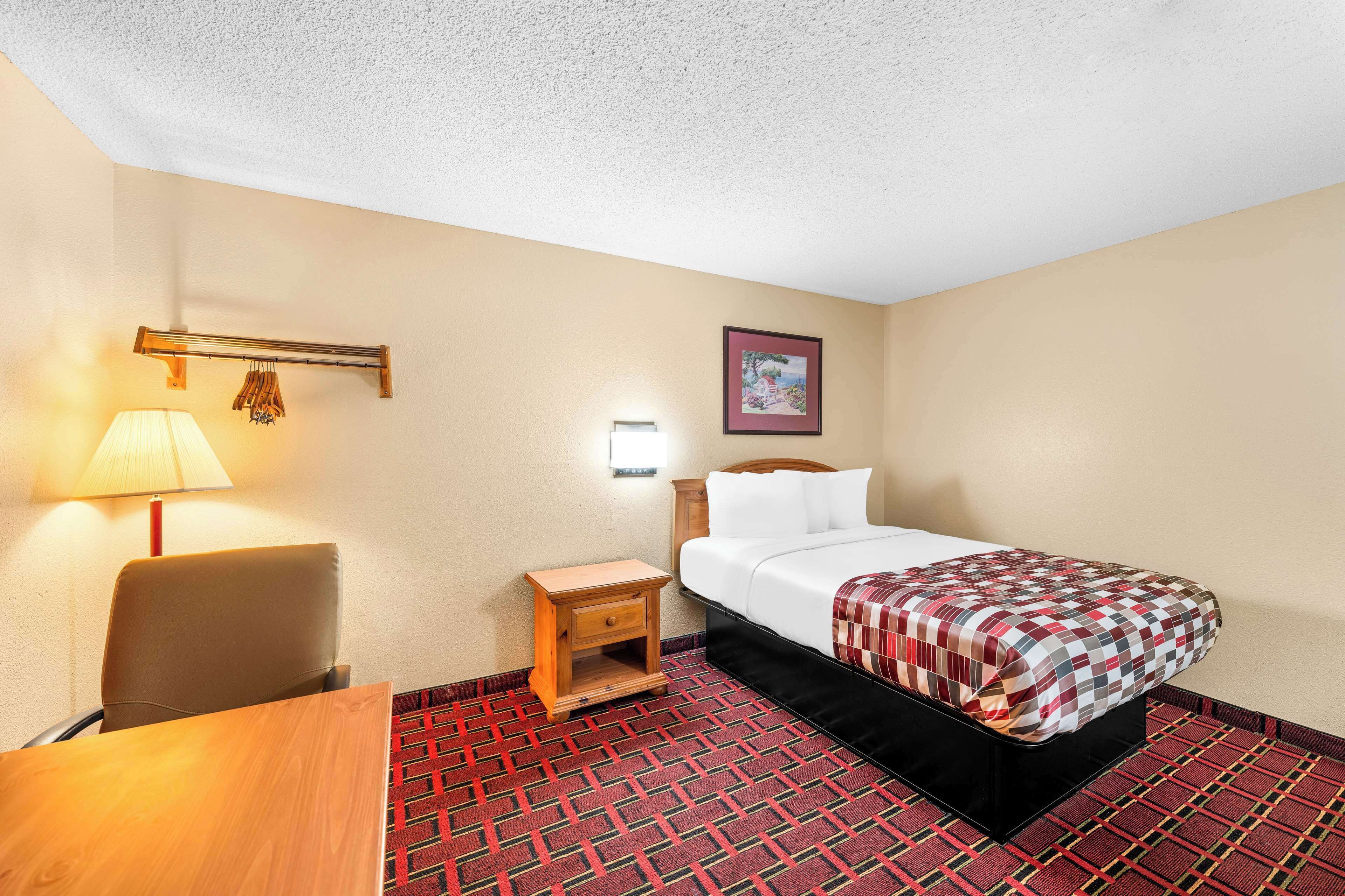 A hotel room with a bed, a desk, a chair, a nightstand, a lamp, and a wall-mounted shelf.