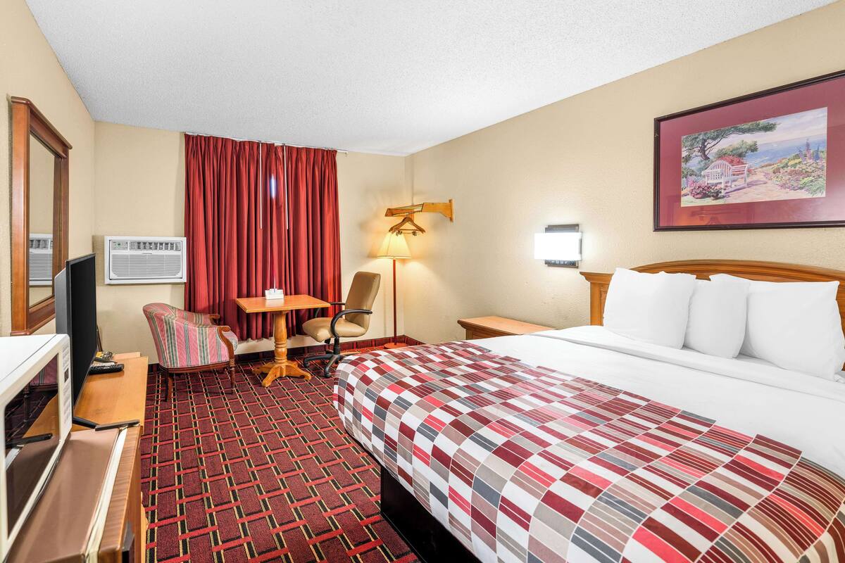 A hotel room with a bed, a desk, a chair, a TV, and a painting on the wall.