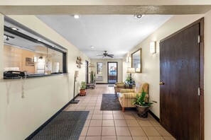 Um hall de hotel com balcão de recepção, ventilador de teto, área de estar com cadeiras e uma grande porta de madeira.