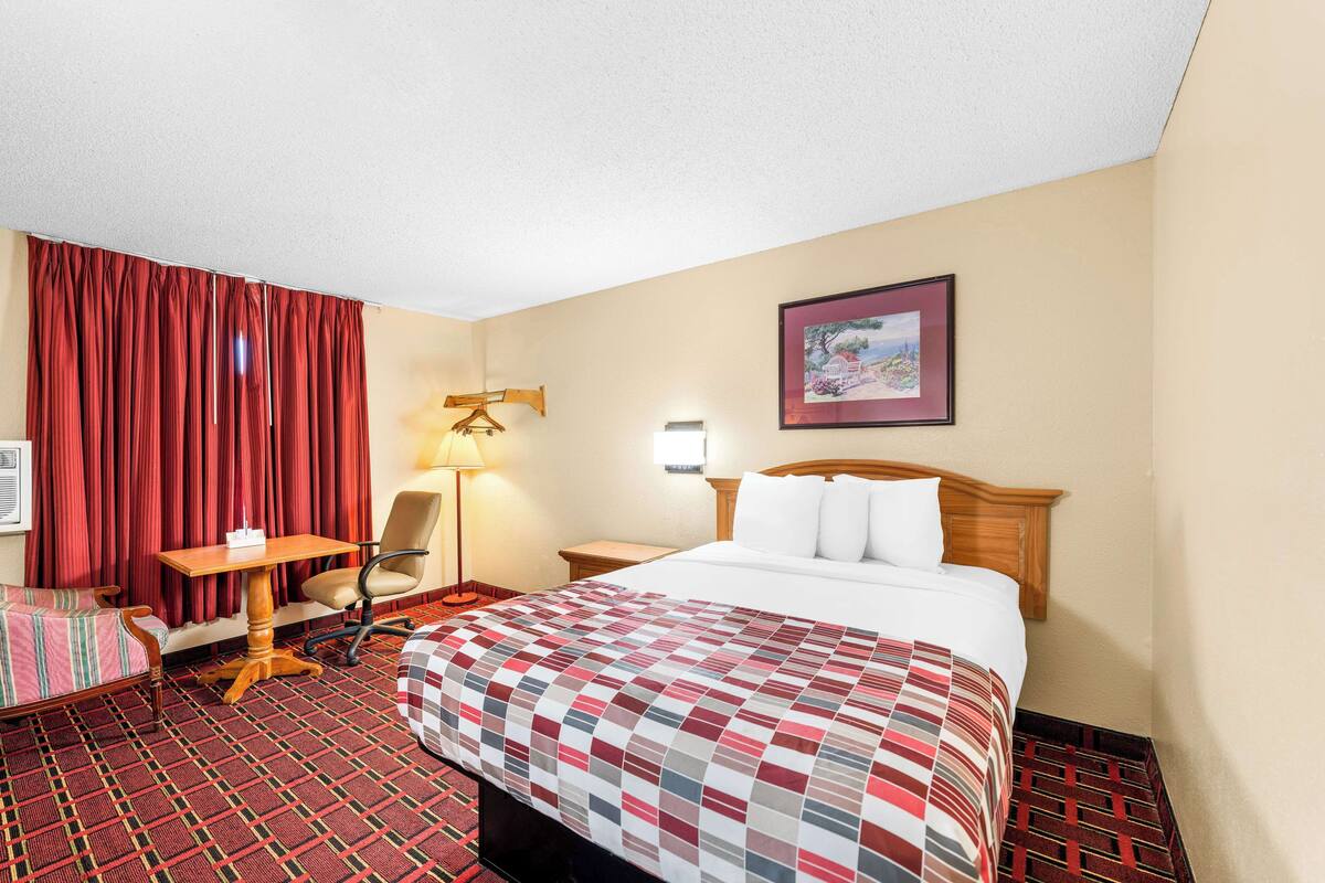 A hotel room with a bed, a desk, a chair, a lamp, and a framed picture on the wall.
