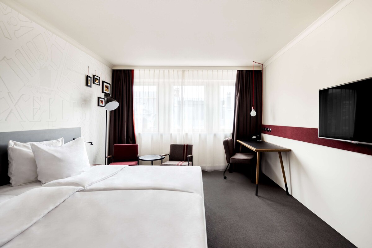 Ein Hotelzimmer mit Bett, Schreibtisch, Sessel, Fernseher und einem Fenster mit Vorhängen.