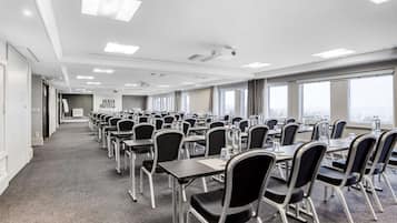 Une salle de conférence avec des rangées de tables et de chaises, de grandes fenêtres et une enseigne indiquant « OENTA HOTELS ».